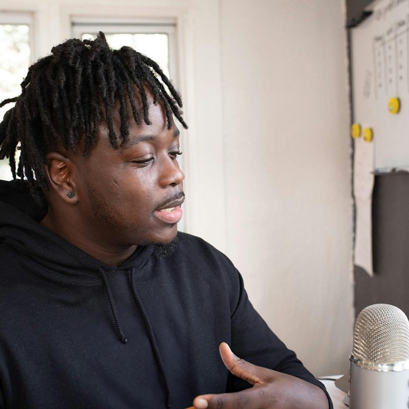 A young man with textured hair styled in loose dreadlocks is sitting at a table, wearing a black hoodie. He is animatedly speaking, gesturing with his hands, and is positioned next to a microphone. The background features a bright, airy room with large windows and a white wall with colorful buttons and notes. The atmosphere conveys a casual yet engaging discussion setting.