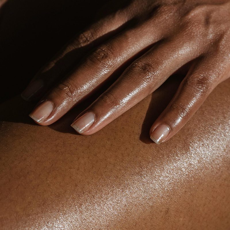 A close-up of a hand resting gently on a smooth, glistening skin surface. The hand features neat, light-colored fingernails, while the skin below is a rich brown tone, radiant with a soft sheen. The background is softly blurred, emphasizing the textures of the skin and the delicate fingers. Natural light enhances the overall warmth of the image.